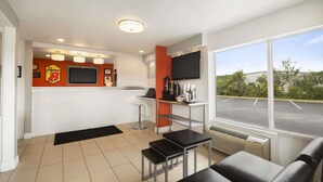 Una habitación de hotel moderna con un ventanal grande, un televisor de pantalla plana, una kitchenette pequeña y un área de descanso con un sofá negro y una mesita.