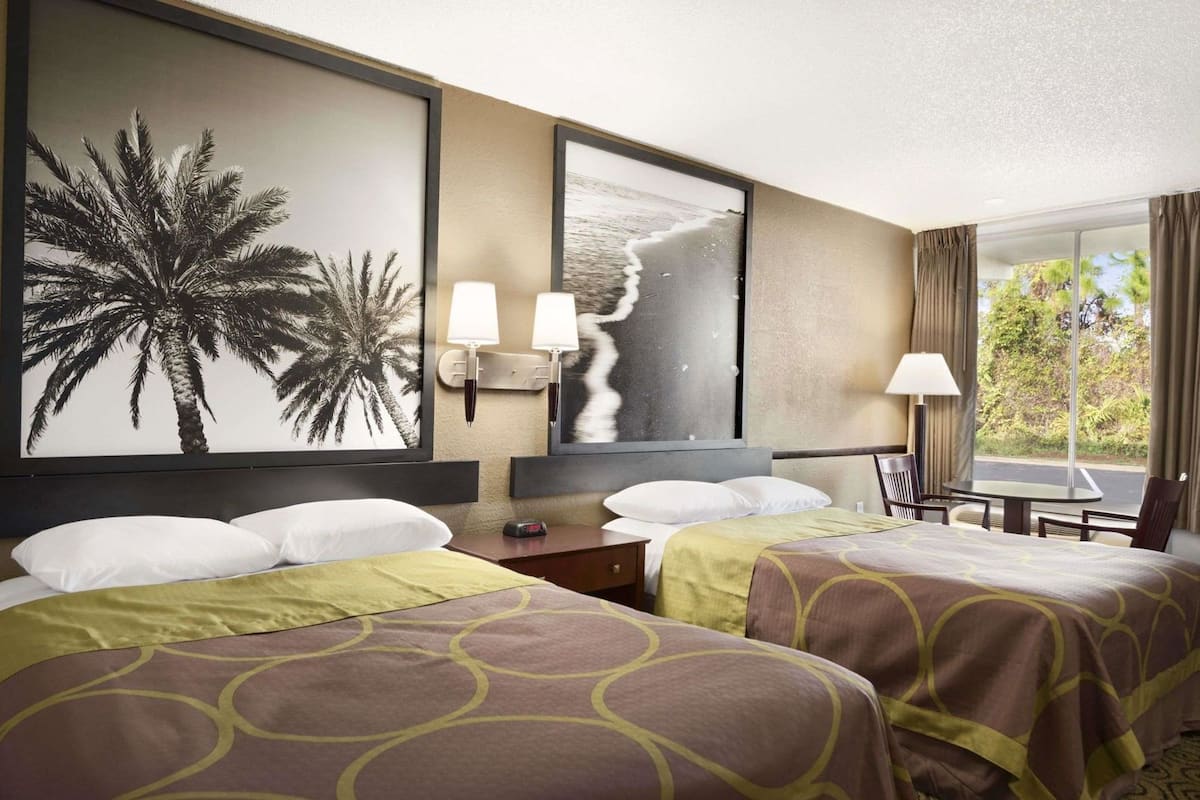 Habitación de hotel con dos camas, un ventanal con cortinas, una mesita de noche, una lámpara y decoración en la pared que muestra palmeras y una playa.