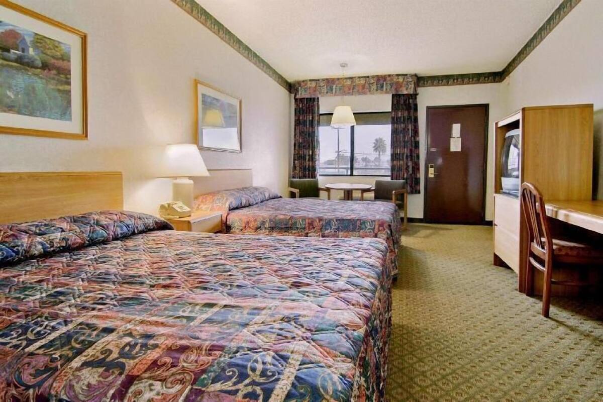 Habitación de hotel con dos camas, un escritorio, una silla, una lámpara, un cuadro y una ventana con cortinas.