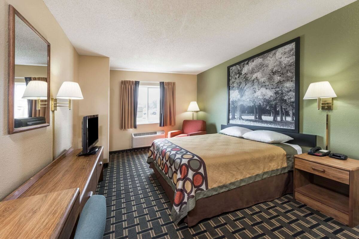 Habitación de hotel con cama, escritorio, silla, televisor, ventana con cortinas y un cuadro grande en la pared.