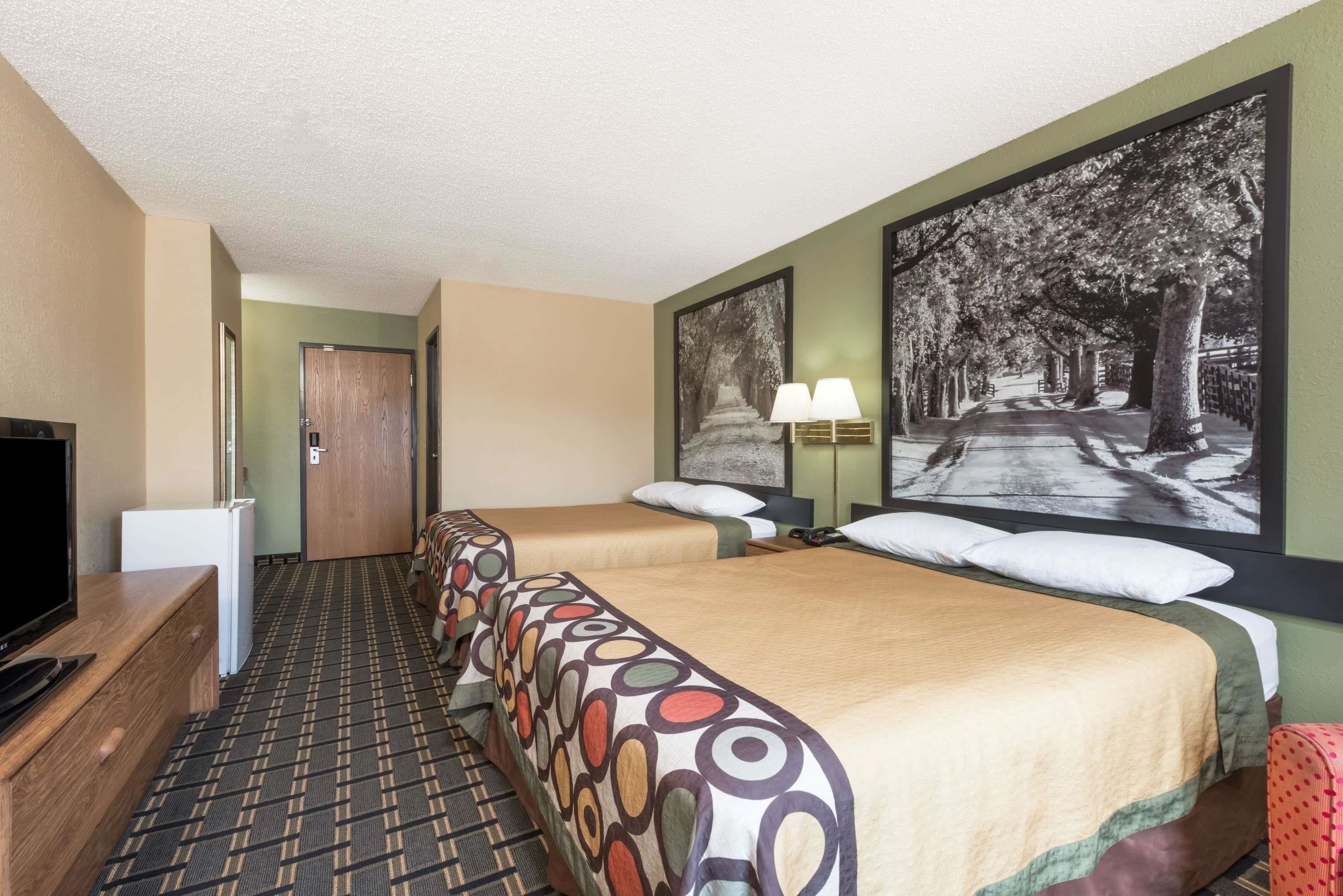Habitación de hotel con dos camas, televisor, escritorio y un cuadro grande con vista a una calle arbolada.