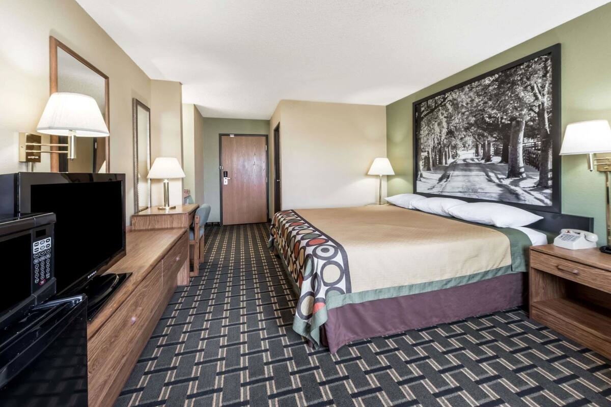 Habitación de hotel con cama, dos mesitas de noche con lámparas, escritorio con televisor, alfombra estampada y puerta que da a otra habitación.