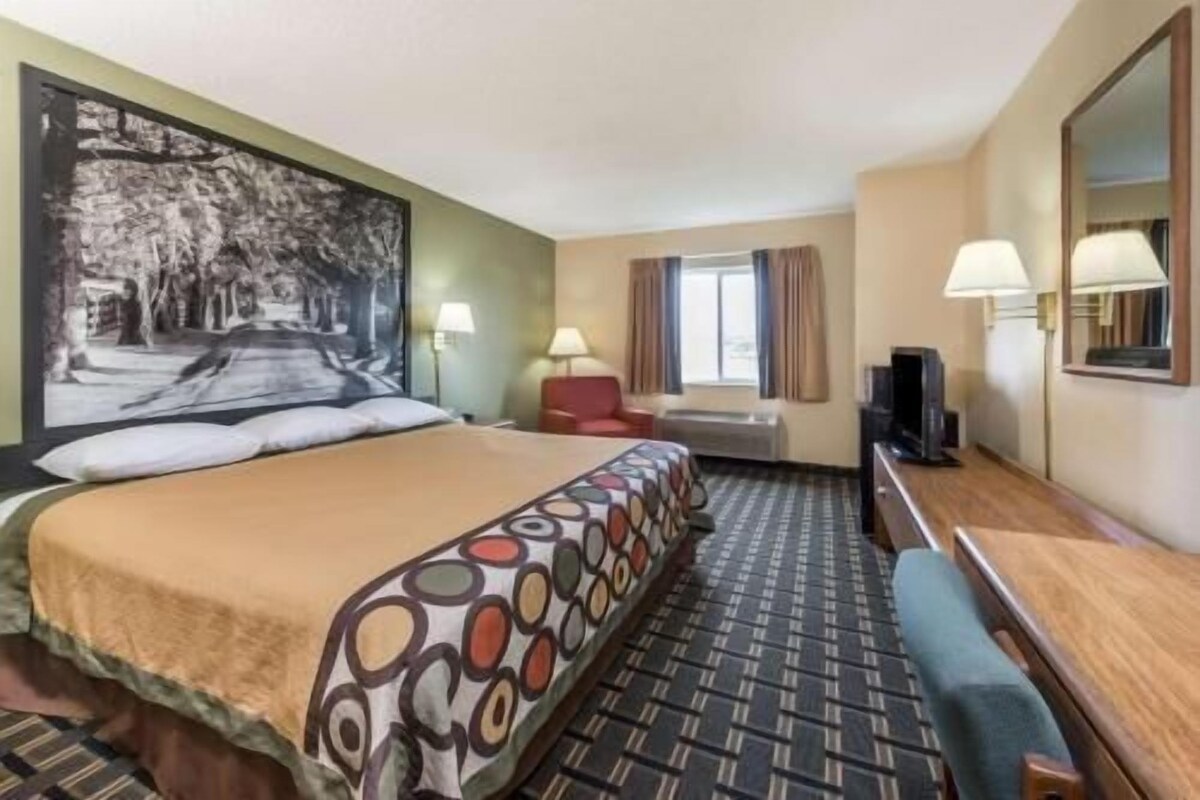 Habitación de hotel con cama, escritorio, televisor, ventana con cortinas y un cuadro en la pared.