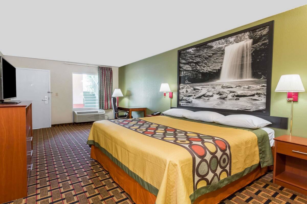 Ein Hotelzimmer mit Bett, Schreibtisch, Stuhl, Fernseher, einem Fenster mit Vorhängen und einer Wandlampe.