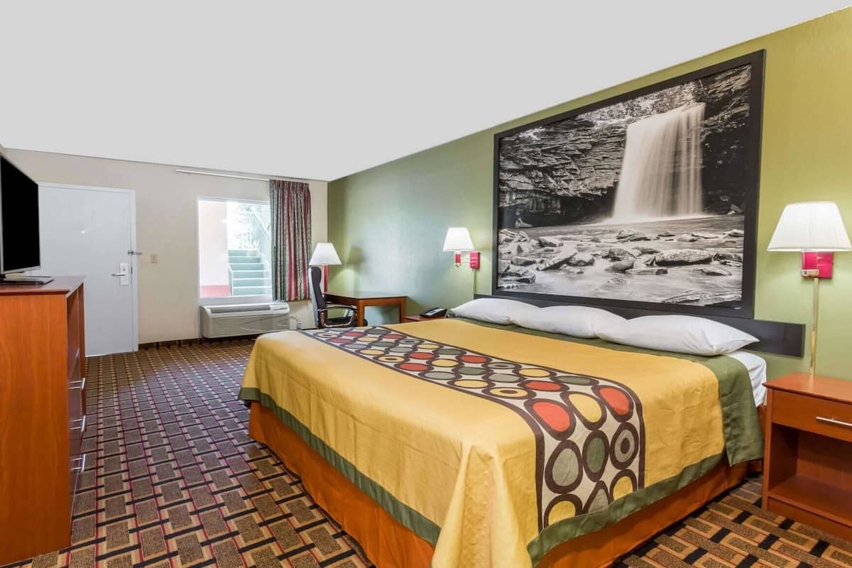 Ein Hotelzimmer mit Bett, Schreibtisch, Fernseher, einem Fenster mit Vorhängen, einer Wandlampe und einem großen Gemälde eines Wasserfalls.