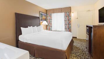 A hotel room with a large bed, a desk with a flat-screen TV, a bathroom with a bathtub, and a patterned carpet.