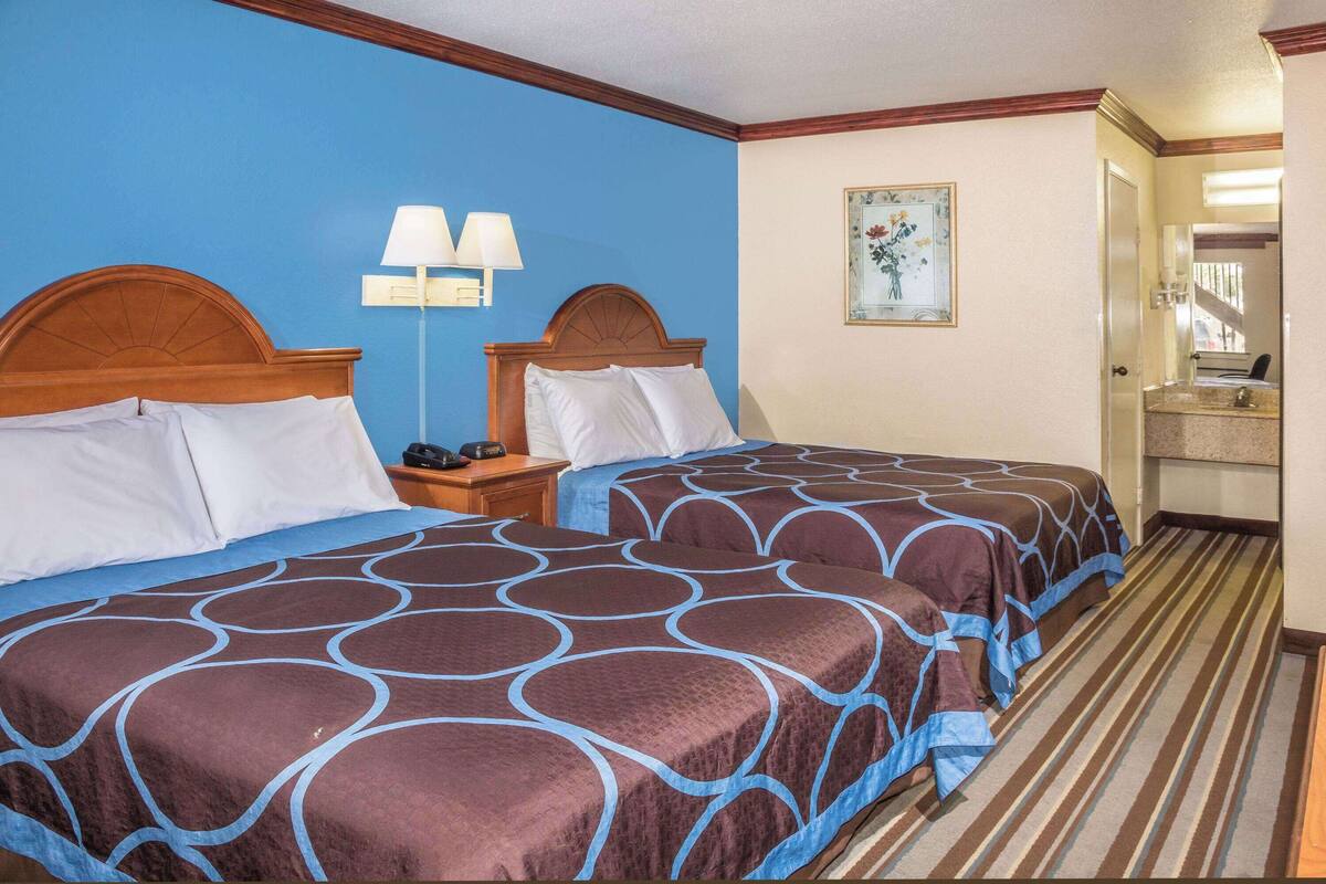 Ein Hotelzimmer mit zwei Betten, einem Nachttisch mit Lampen, einem Bild an der Wand und einem durchgegangenen Tür erkennbaren Badezimmer.