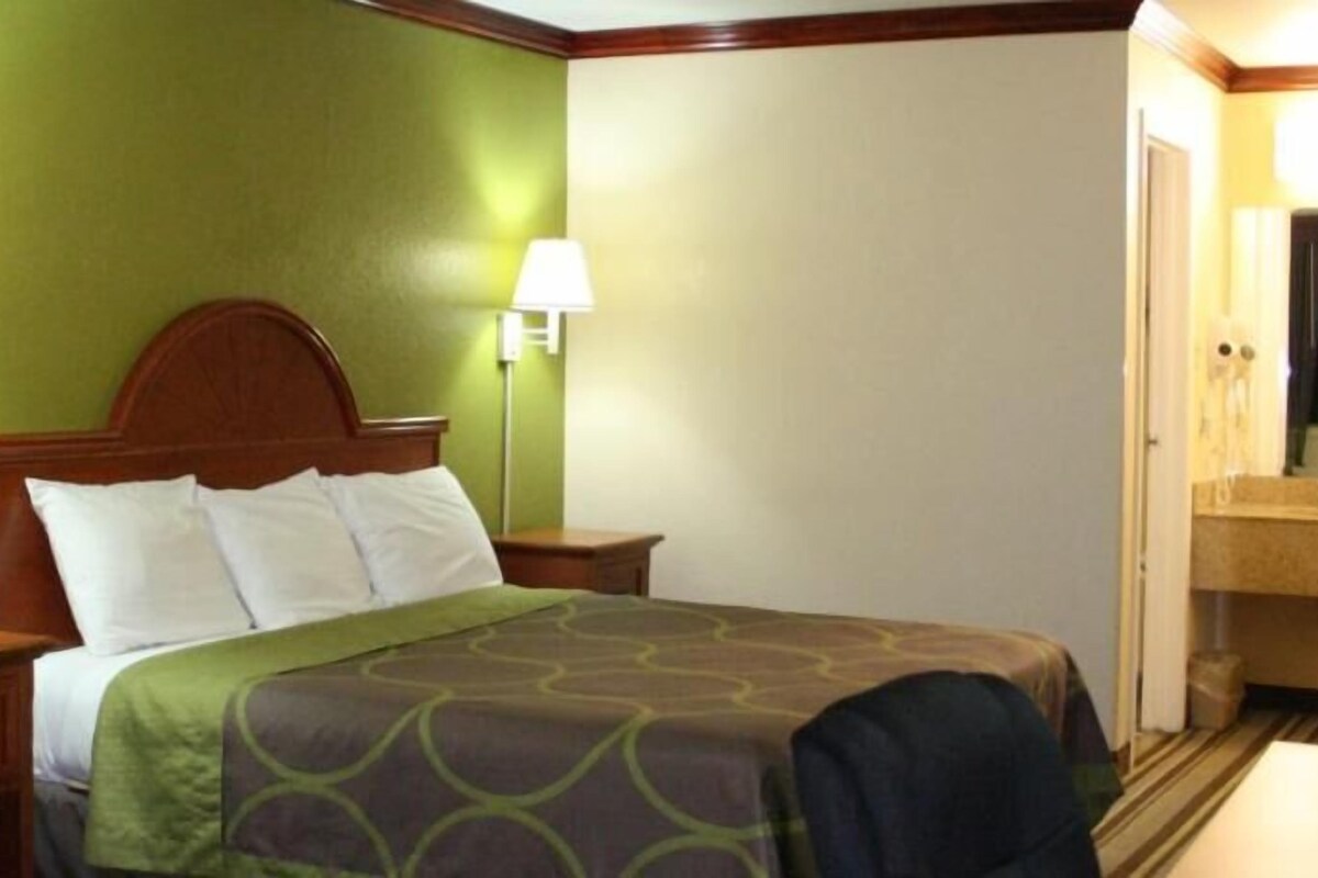 Habitación de hotel con cama, dos lámparas de noche, cabecero de madera, pared verde y baño con lavamanos y espejo.