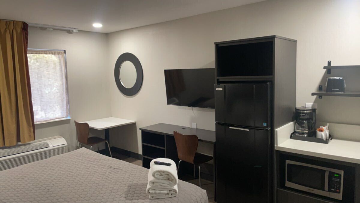 Habitación de hotel con cama, escritorio, silla, televisor, refrigerador, microondas y cafetera.