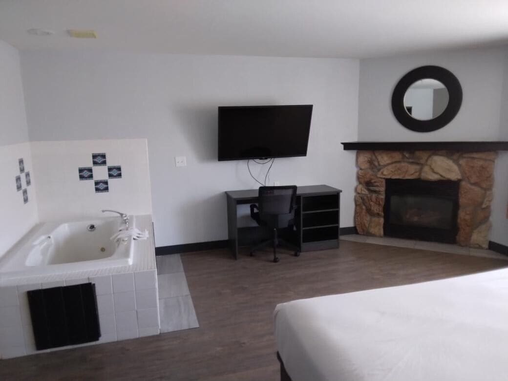 A hotel room with a large bed, a desk with a chair, a television, a fireplace, and a bathtub.