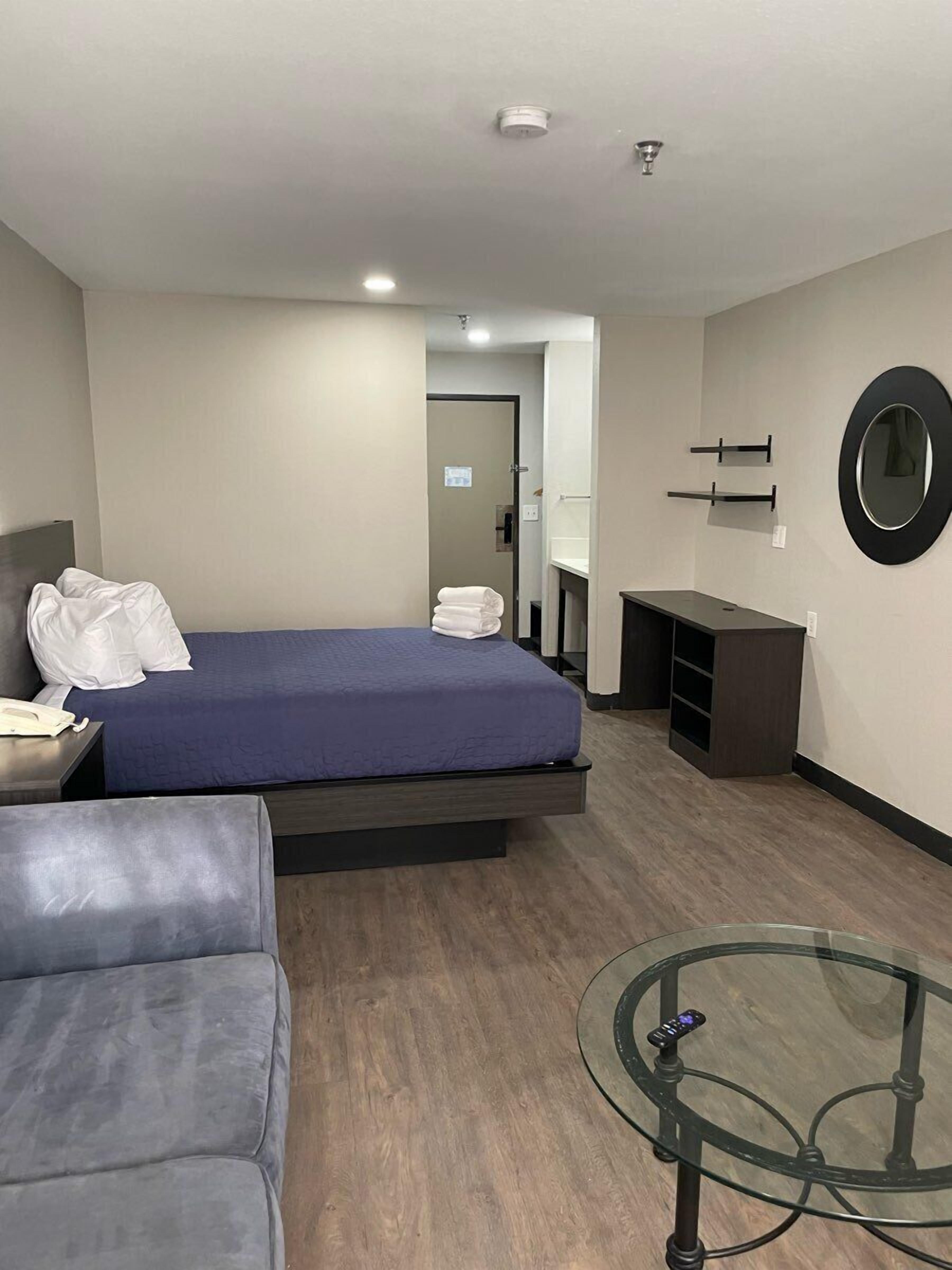 A modern hotel room with a bed, a sofa, a glass coffee table, a desk, and a mirror on the wall.
