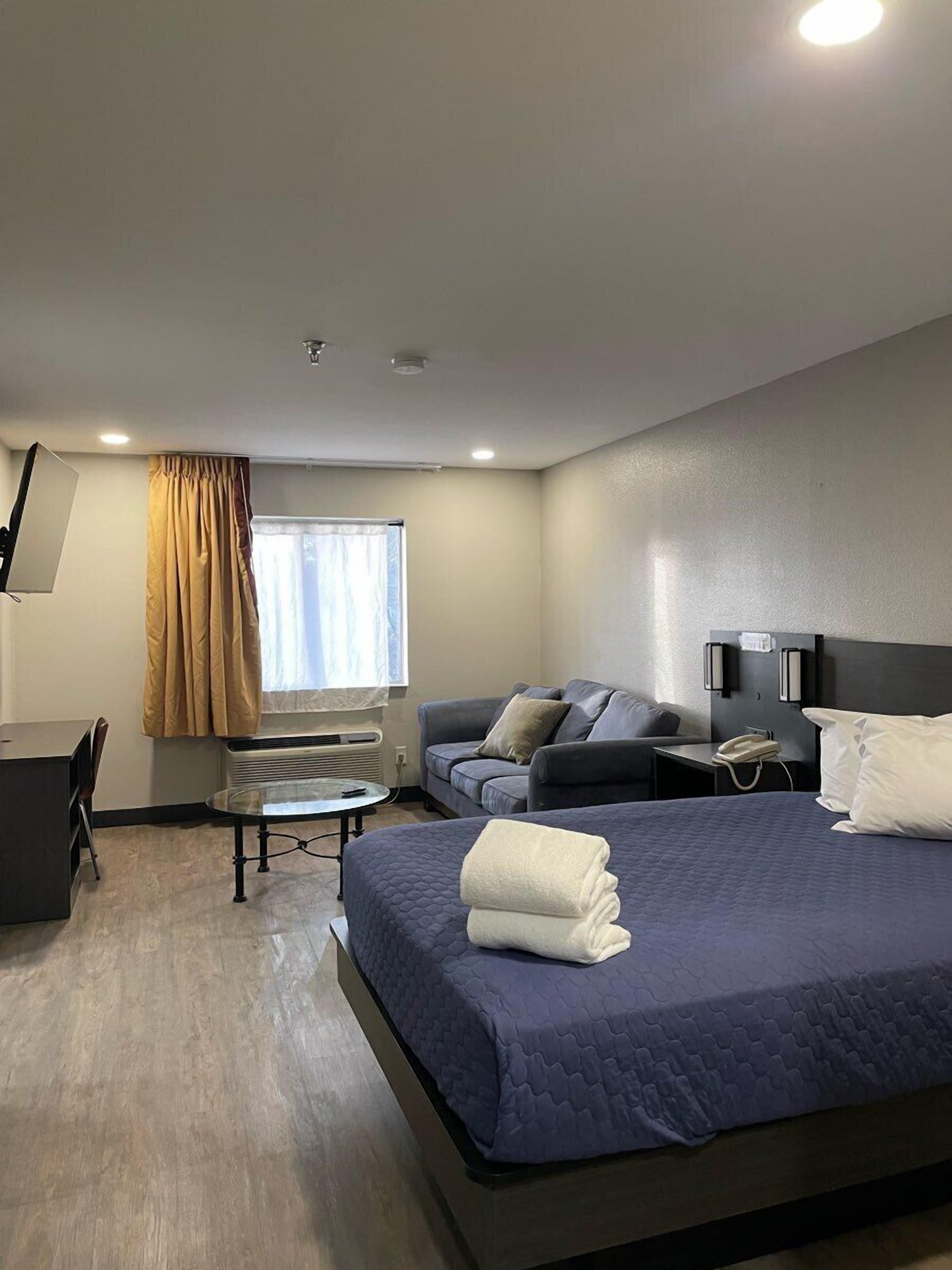 A modern hotel room with a large bed, a desk, a chair, a sofa, and a bedside table with a phone.