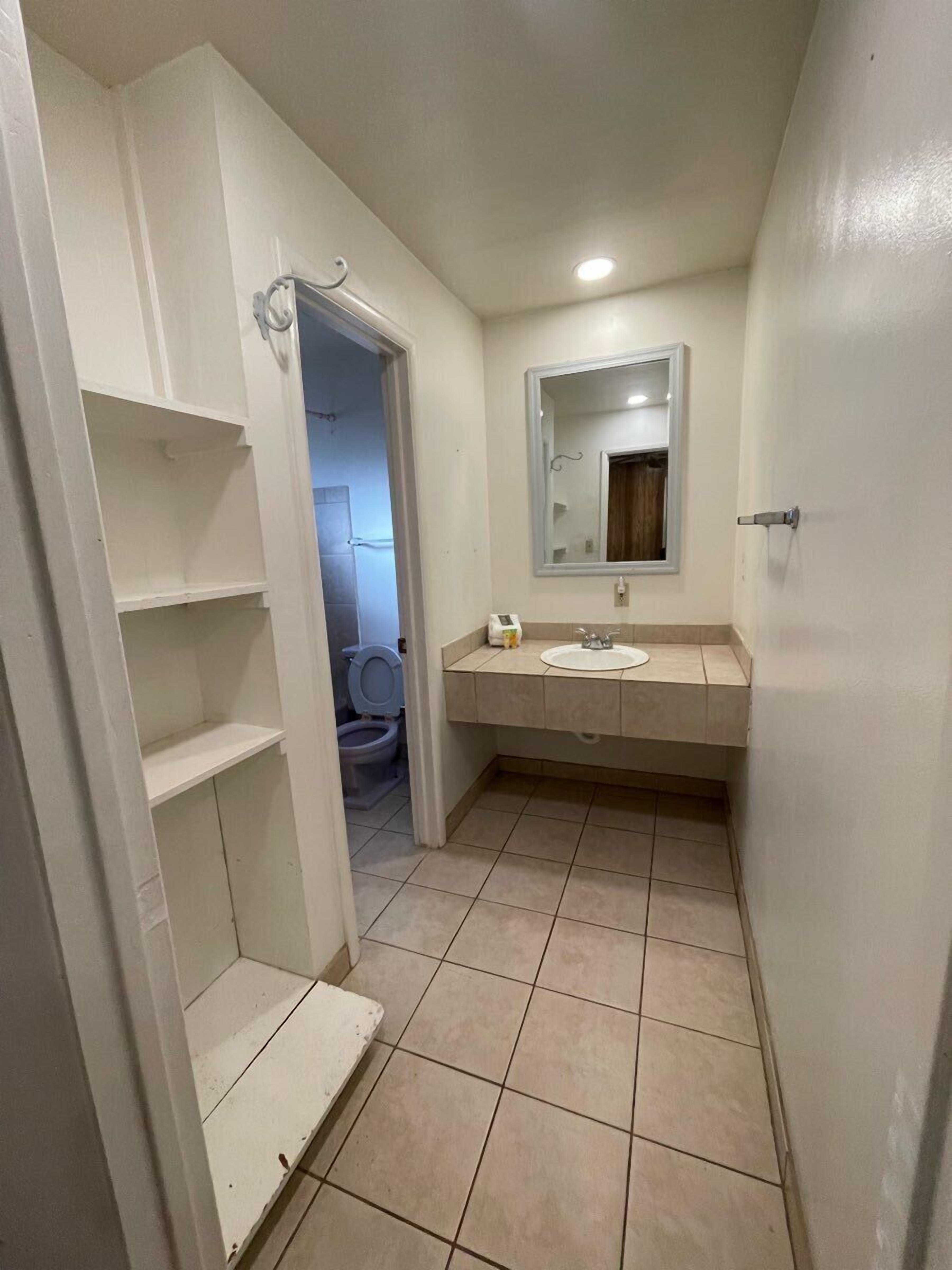 Un baño estrecho con inodoro, lavamanos y espejo. Hay estantes blancos en la pared izquierda y una puerta que da a otra habitación.