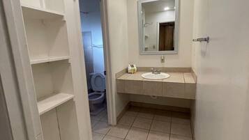 A narrow bathroom with a toilet, a sink, and a mirror. There are white shelves on the left wall and a door leading to another room.