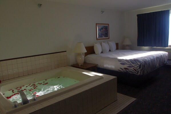 Une chambre d’hôtel avec une grande baignoire remplie d’eau et de pétales de rose, un lit recouvert d’une couvre-lit à motifs, deux lampes de chevet et un tableau encadré au mur.