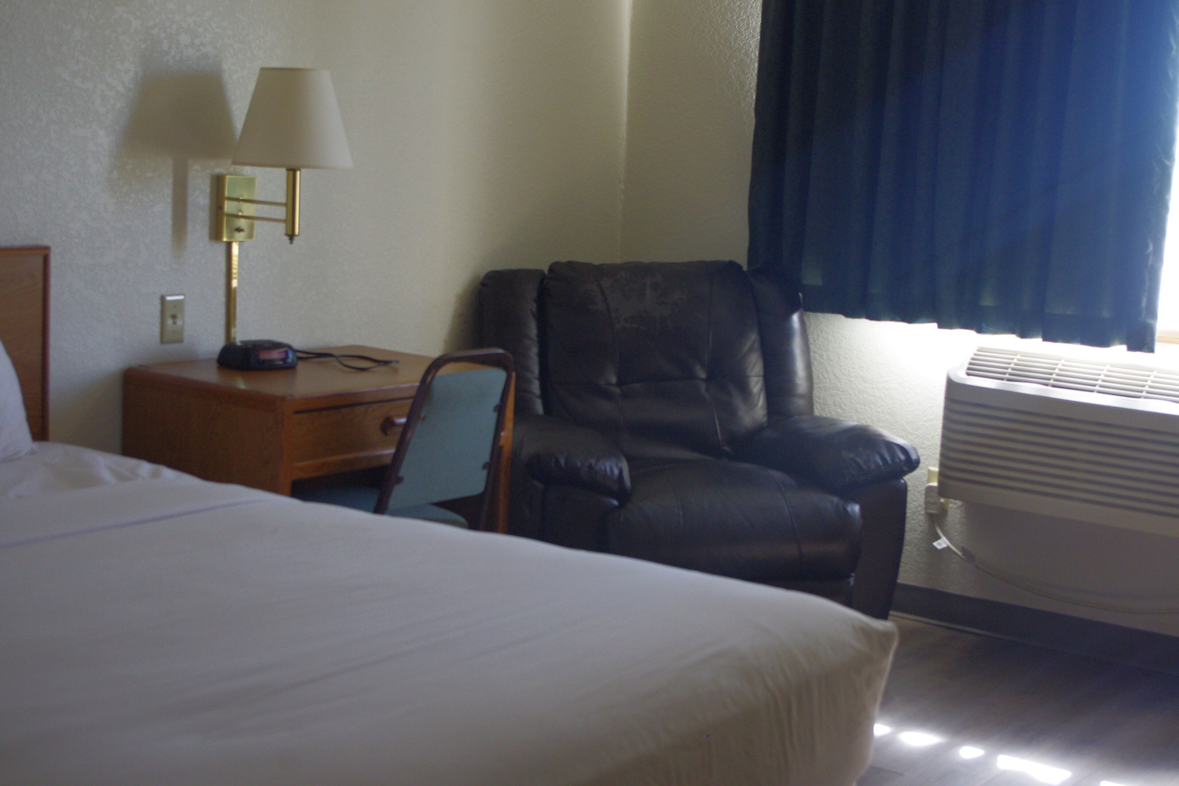A hotel room with a bed, a desk with a lamp, a leather chair, and an air conditioning unit.