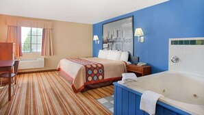 Une chambre d’hôtel comprenant un lit, un bureau, une chaise, une fenêtre avec des rideaux, un grand miroir, une lampe fixée au mur et une baignoire.