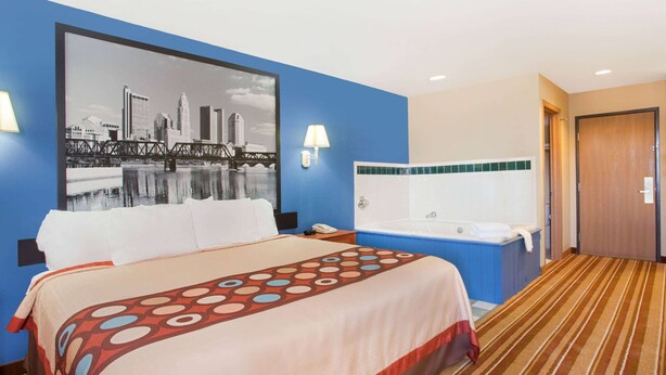 Une chambre d’hôtel avec un grand lit, un tableau représentant un paysage urbain accroché au mur, une baignoire et une porte sur le côté droit.