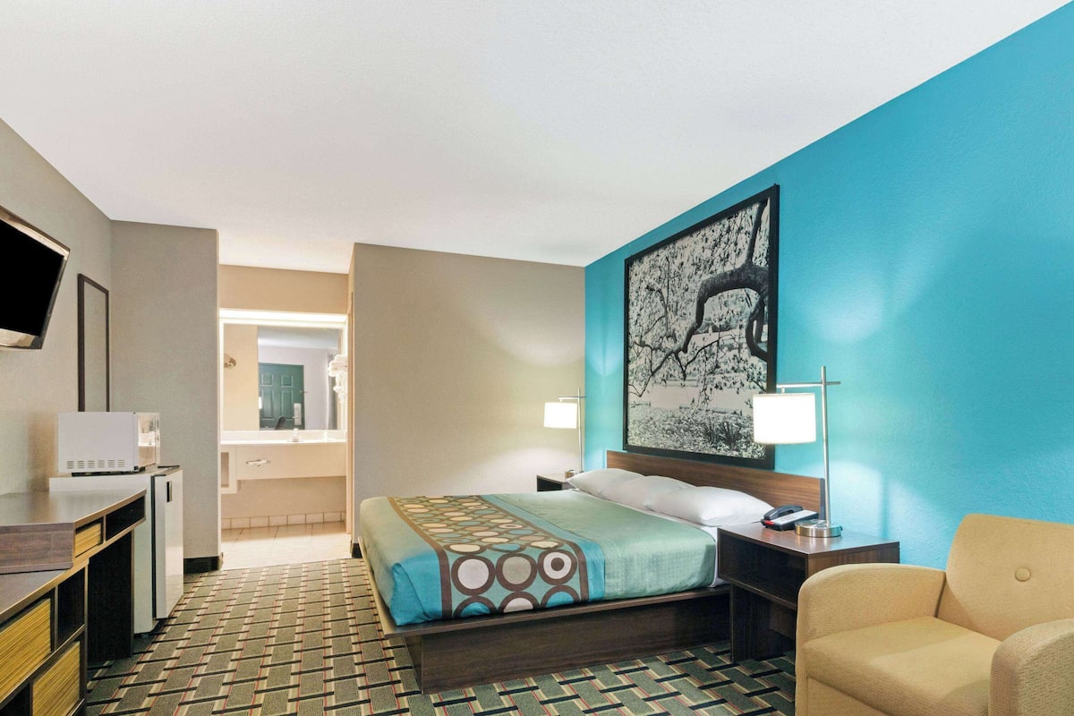 A hotel room with a bed, a desk, a chair, a TV, and a bathroom visible in the background.