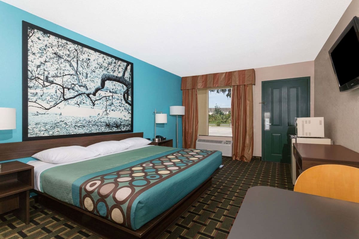 A hotel room with a bed, a desk, a chair, a TV, a window with curtains, and a painting on the wall.