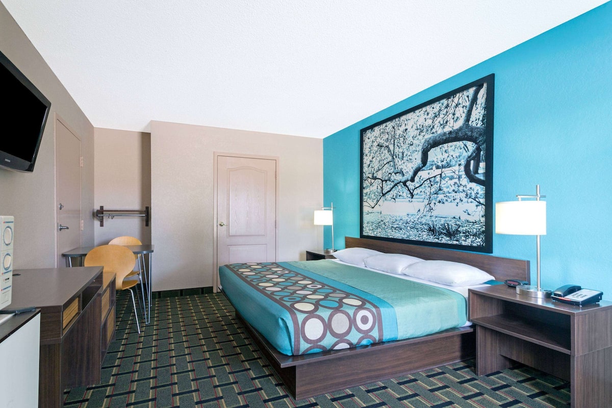 A hotel room with a bed, a desk, a chair, a TV, and a painting on the wall.