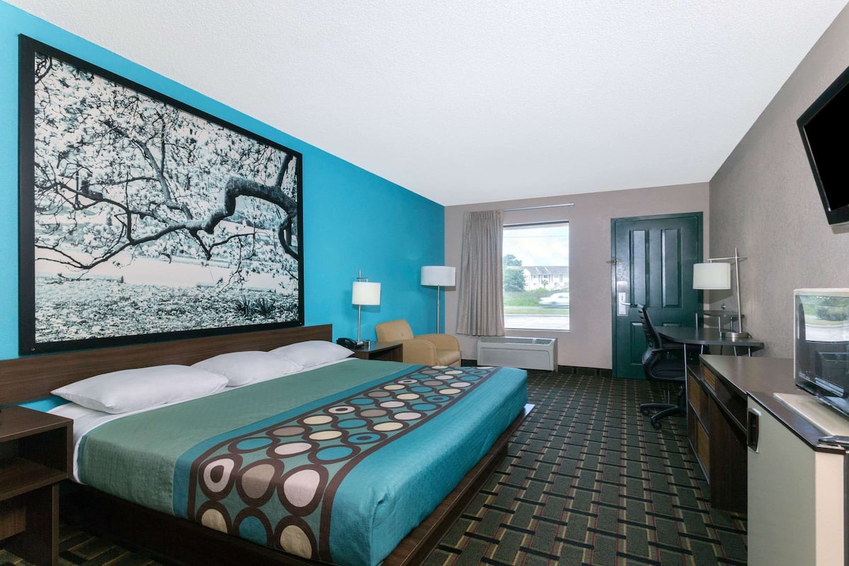 A hotel room with a large bed, a desk with a TV, a chair, a desk lamp, a window with curtains, and a painting on the wall.