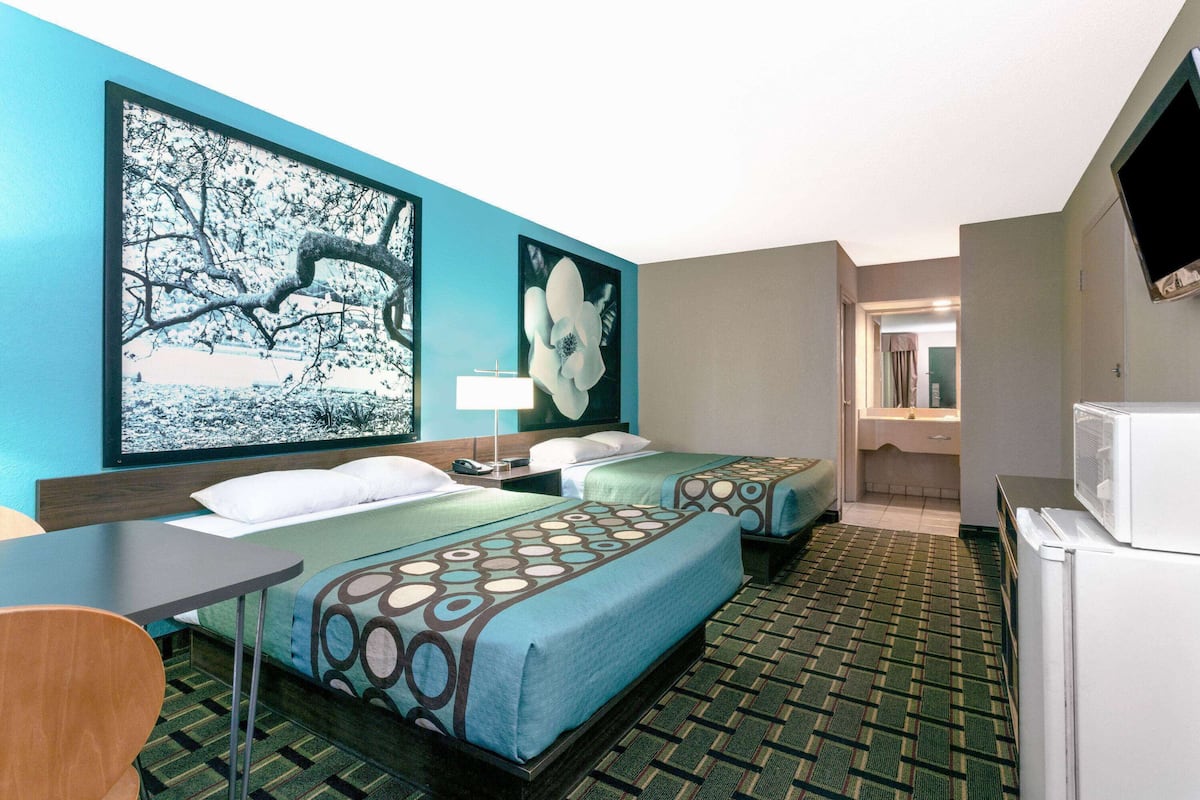 A hotel room with two beds, a desk, a refrigerator, and artwork on the walls.