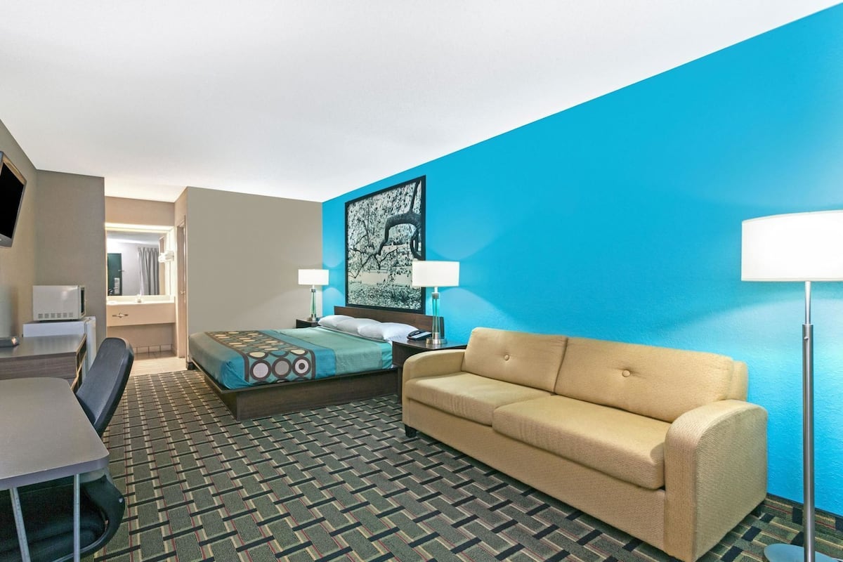 A hotel room with a bed, a sofa, a desk, a chair, a TV, and a painting on the wall.