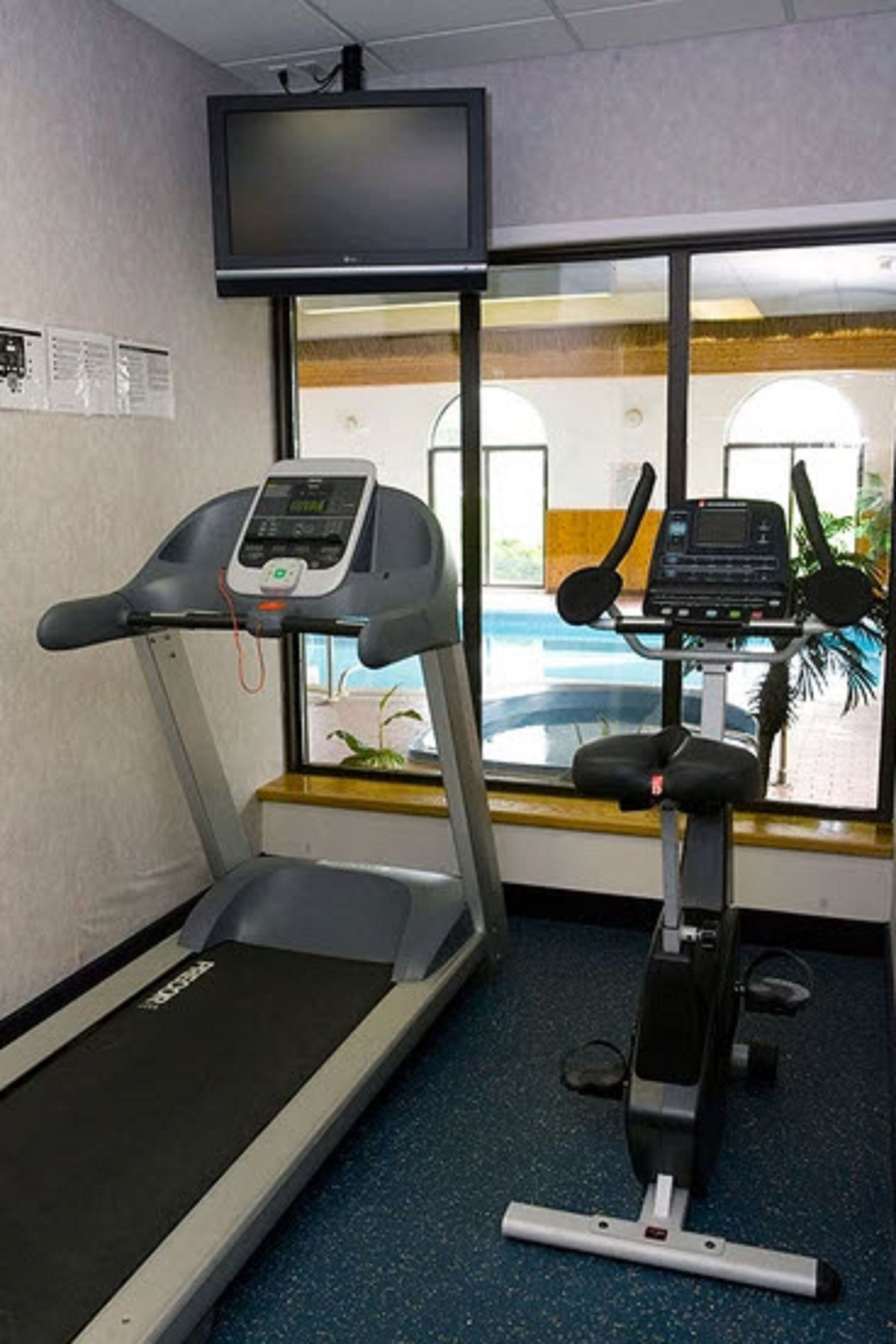 Un gimnasio con cintas de correr y una máquina elíptica, un televisor montado en la pared y una ventana con vistas al exterior.