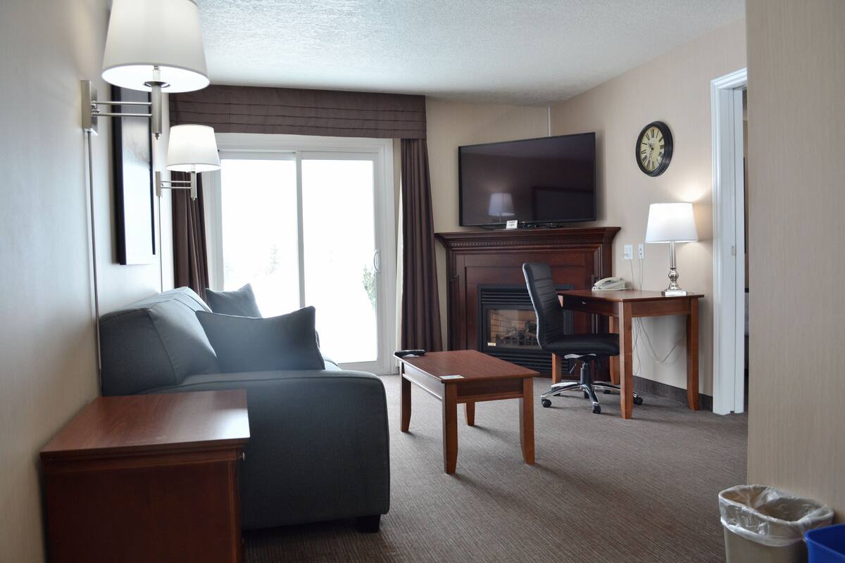 A hotel room with a sofa, a wooden table, a desk with a lamp, a television, and a fireplace.