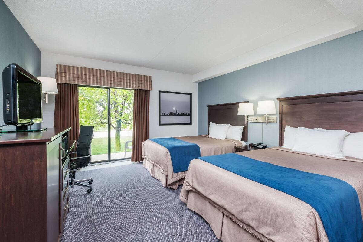 A hotel room with two beds, a desk with a television, a chair, and a window with a view of greenery.