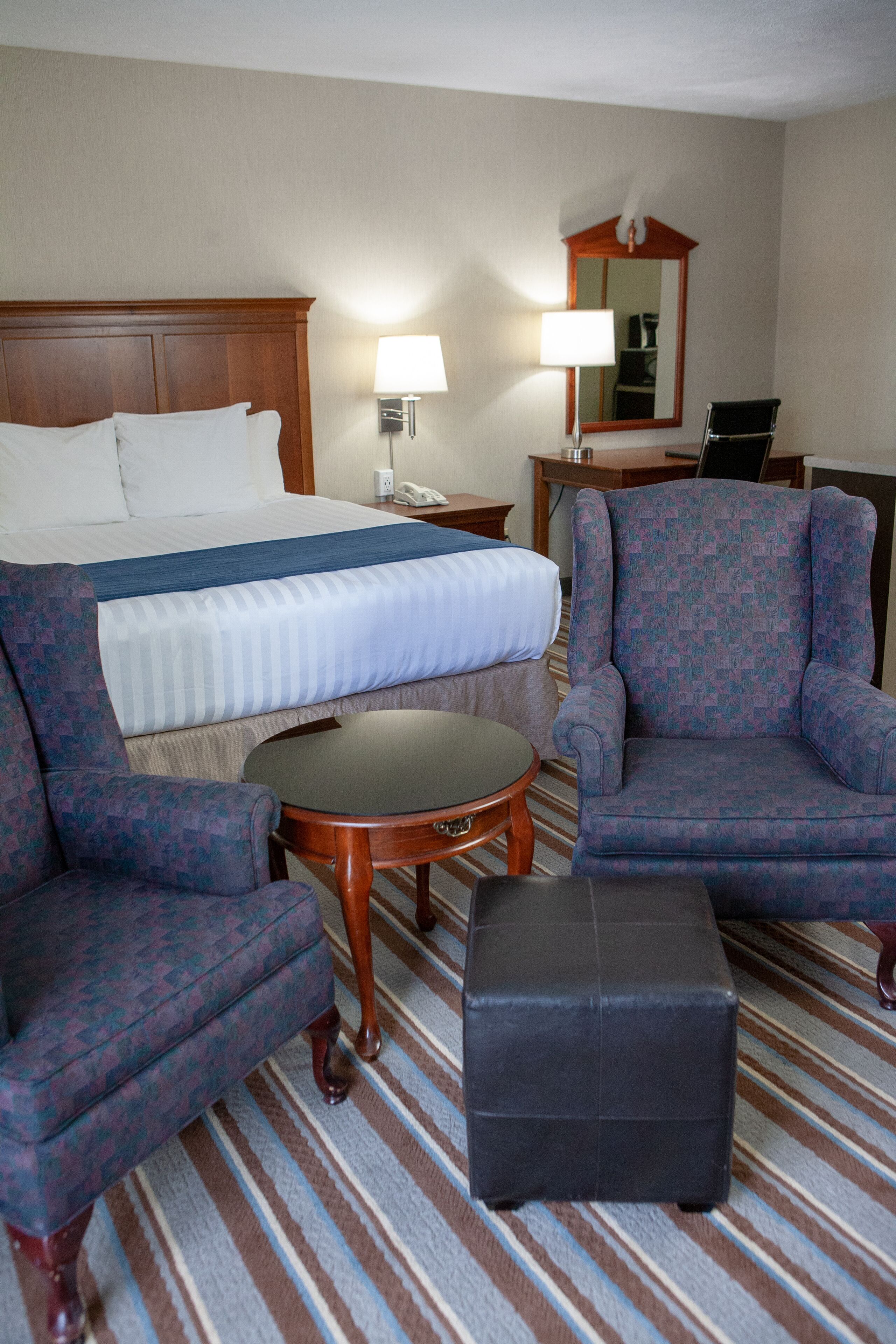 Quarto de hotel com uma cama, duas poltronas, uma mesa pequena, uma luminária, um espelho e uma escrivaninha.