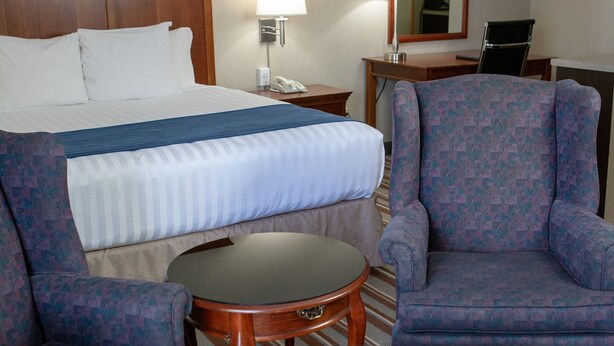 A hotel room with a bed, two armchairs, a small table, a lamp, a mirror, and a desk.