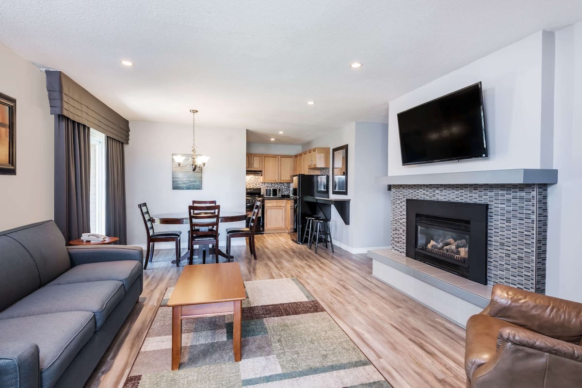 A modern living room with a sofa, coffee table, and fireplace. There is a dining area with chairs and a table. The kitchen is visible in the background with wooden cabinets and a TV mounted above the counter.