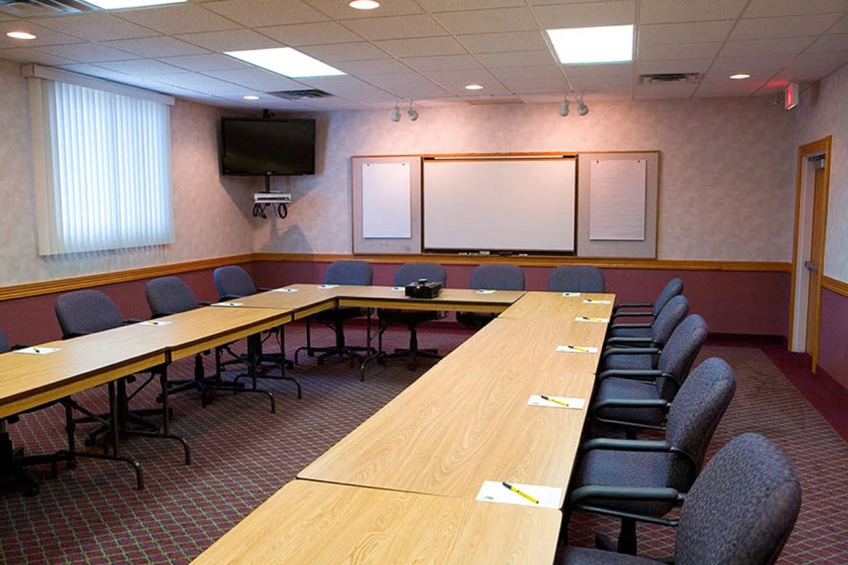 Un salón de conferencias con una larga mesa, sillas, un proyector, una pantalla y un televisor en la pared.