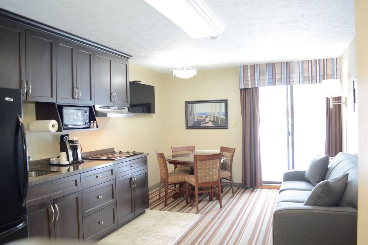 A compact kitchen with dark cabinets, a microwave, and a coffee maker. There is a dining area with a table and chairs, and a gray sofa with a view of the outdoors.