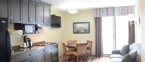A compact kitchen with dark cabinets, a microwave, and a coffee maker. There is a dining area with a table and chairs, and a gray sofa with a view of the outdoors.