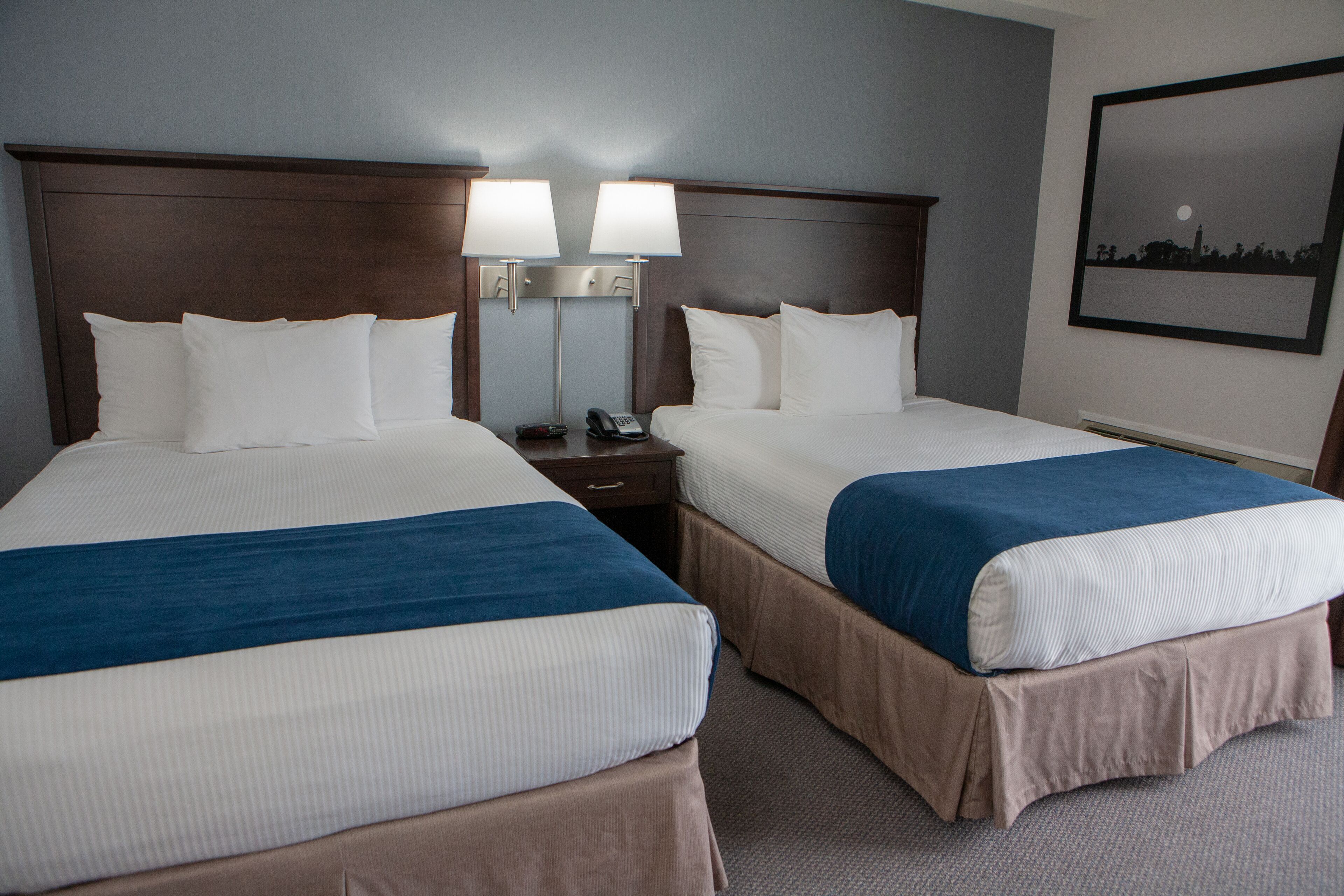 A hotel room with two beds, each with a blue and white bedspread, a nightstand with a phone, and a wall-mounted lamp.
