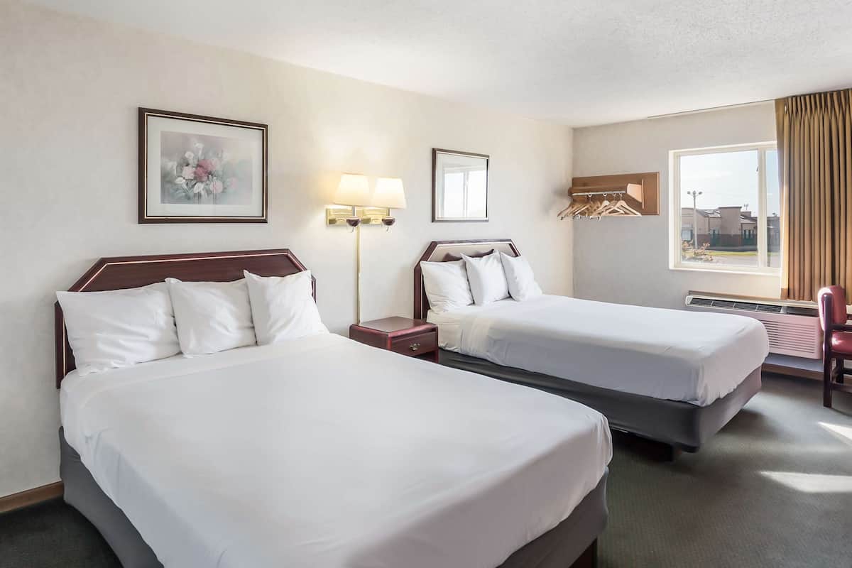 A hotel room with two beds, a nightstand, a lamp, a framed picture, a window with curtains, and a chair.