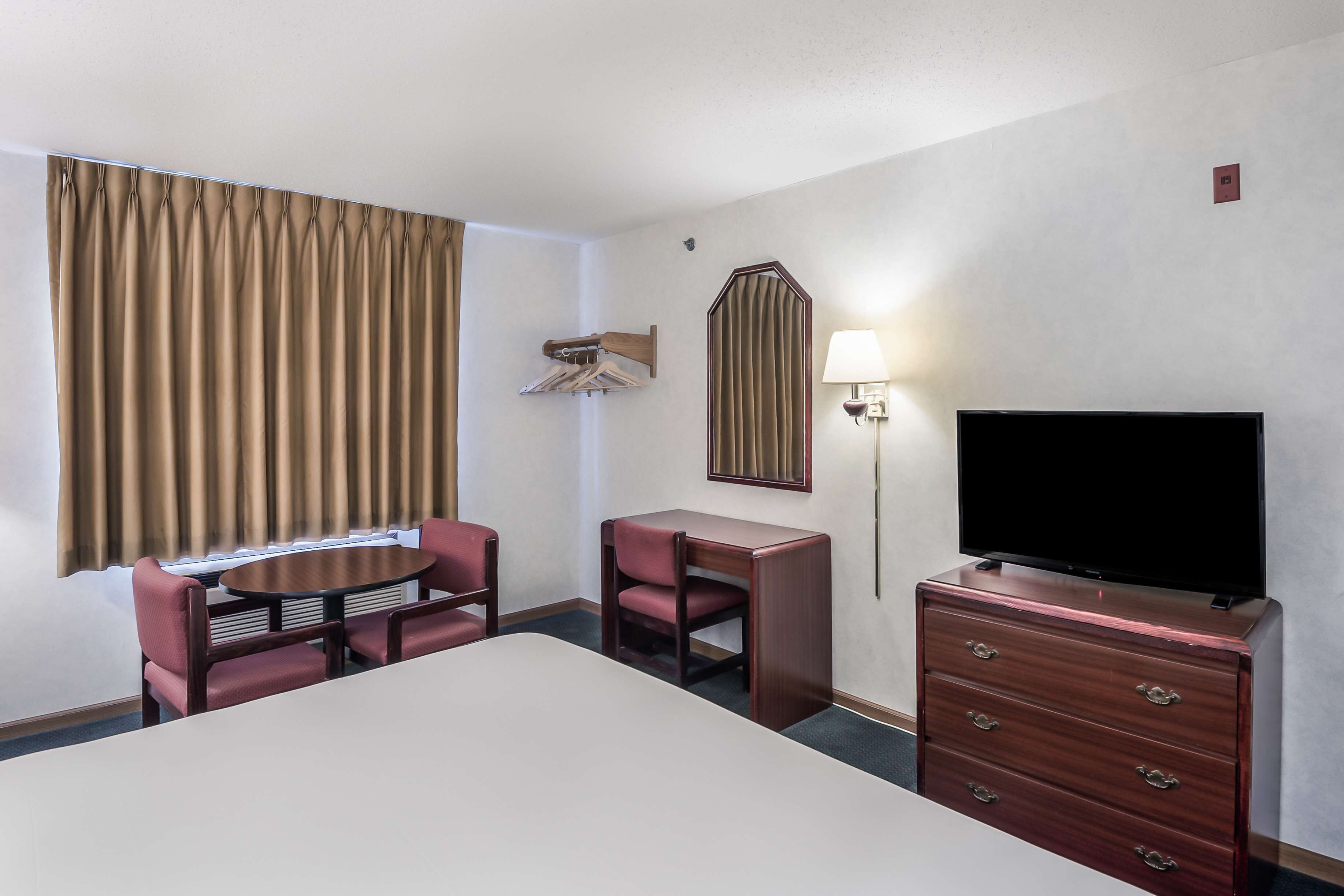A hotel room with a bed, a desk with a chair, a TV on a dresser, and a window with curtains.