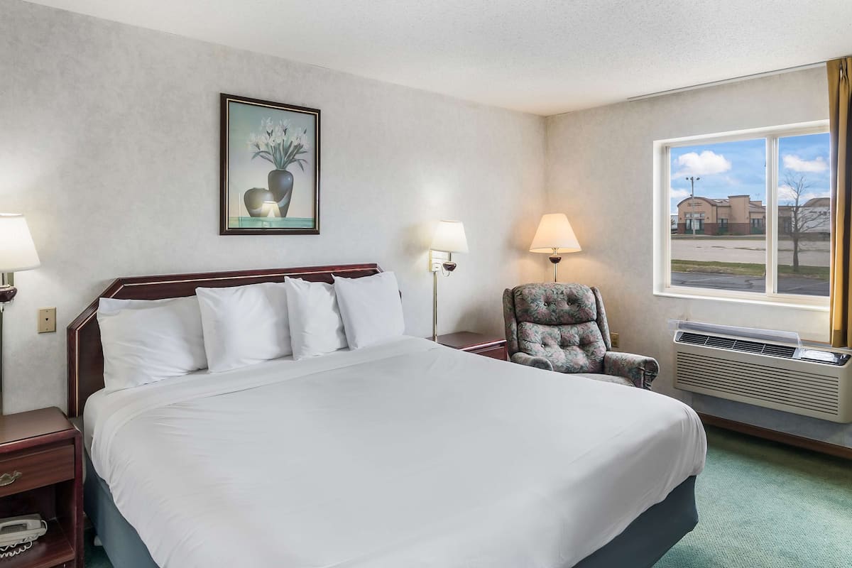 A hotel room with a large bed, two bedside tables, a chair, a window with a view, and a wall-mounted lamp.
