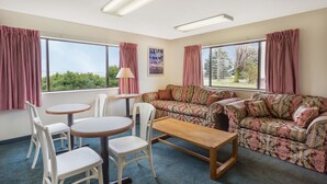 A room with a floral sofa, a wooden table, and white chairs. There are large windows with curtains, a poster on the wall, and a lamp on a side table. - Super 8 by Wyndham Hebron Lowell Area (Hebron)