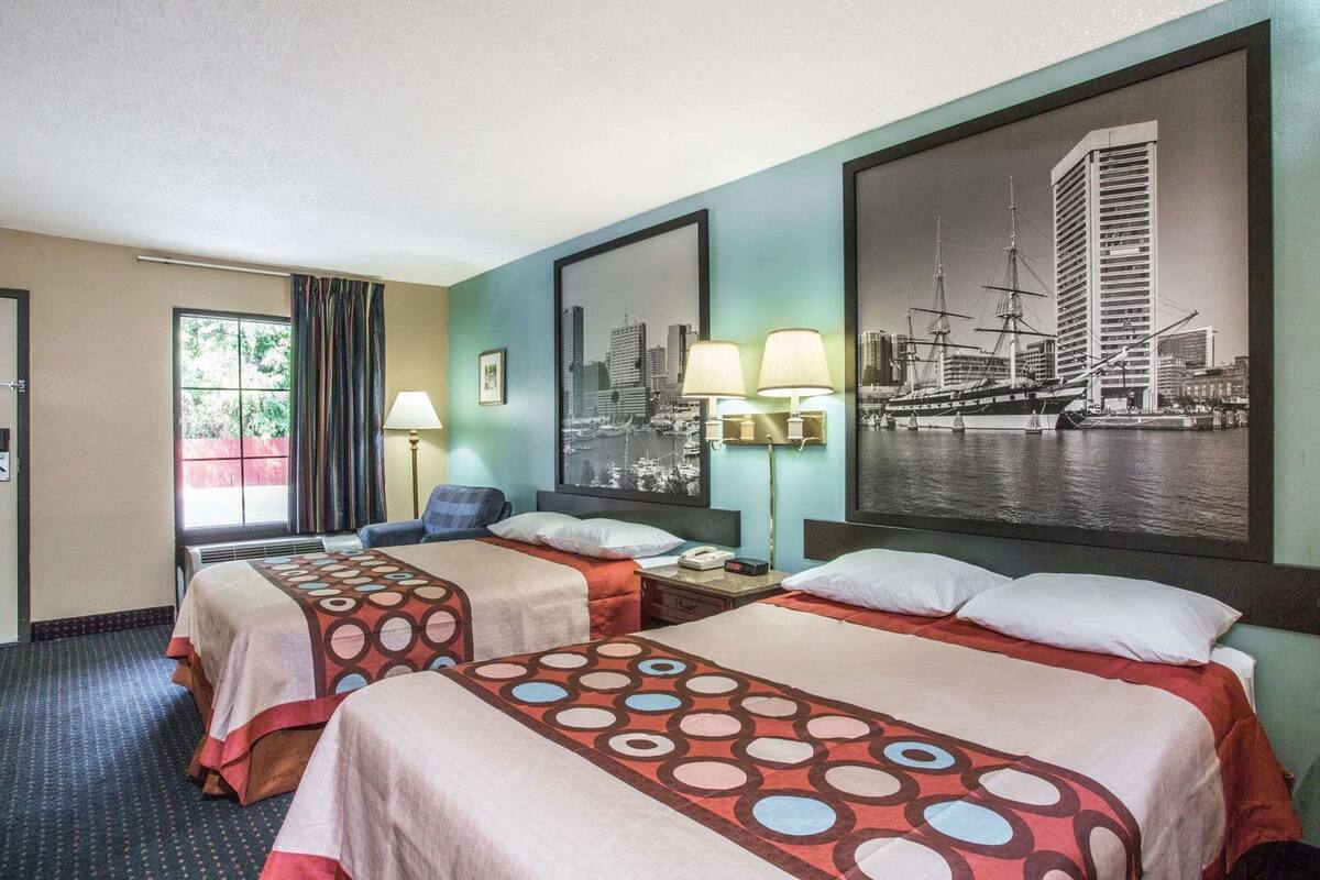 Habitación de hotel con dos camas, una ventana con cortinas, un cuadro de un paisaje urbano y dos lámparas de pared.