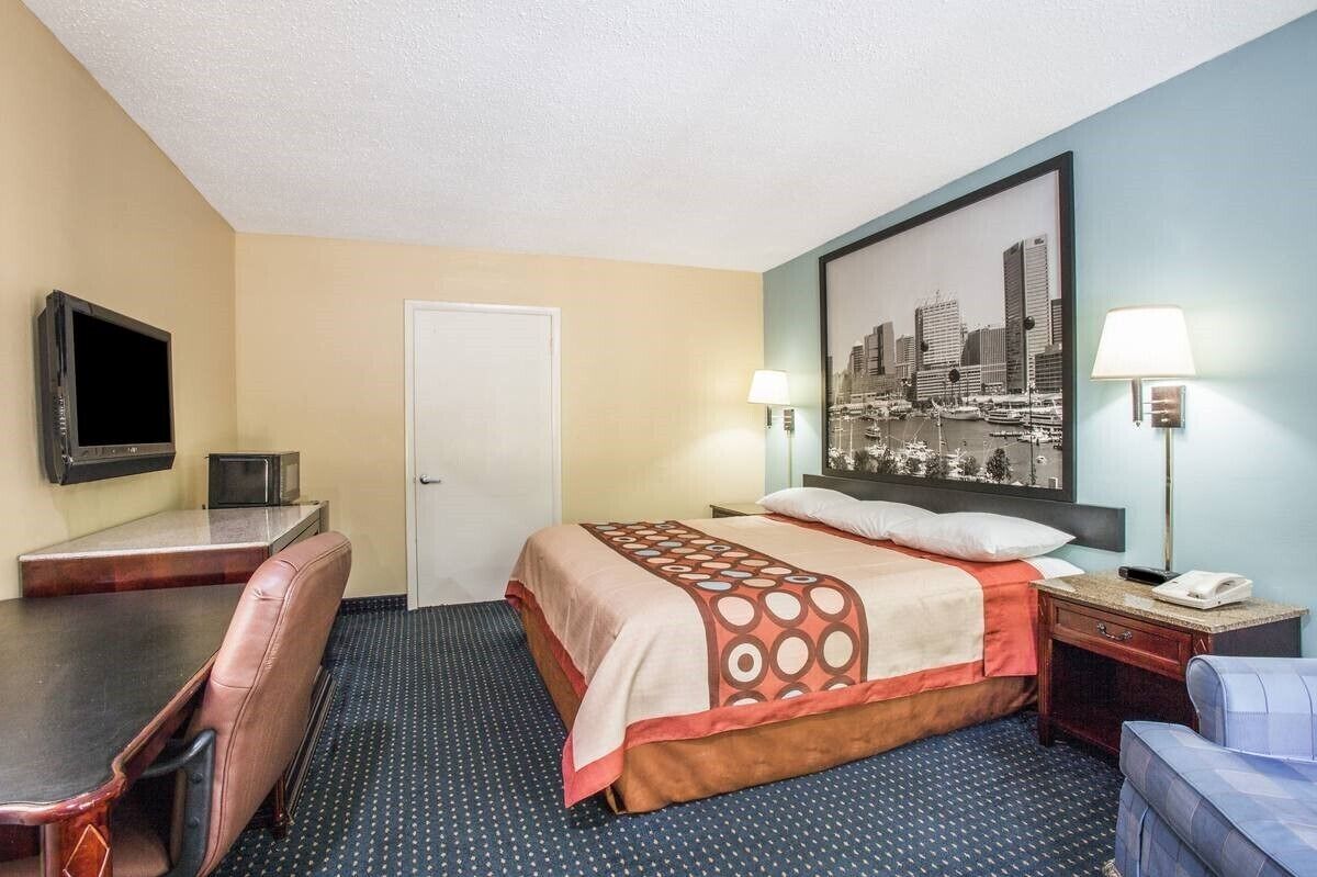 A hotel room with a bed, a desk, a chair, a TV, and a cityscape painting on the wall.