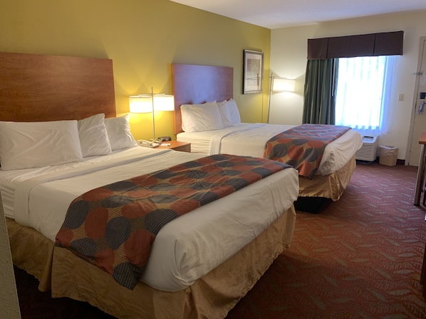 A hotel room with two beds, a wooden headboard, a bedside table with a lamp, a framed picture, and a window with curtains.