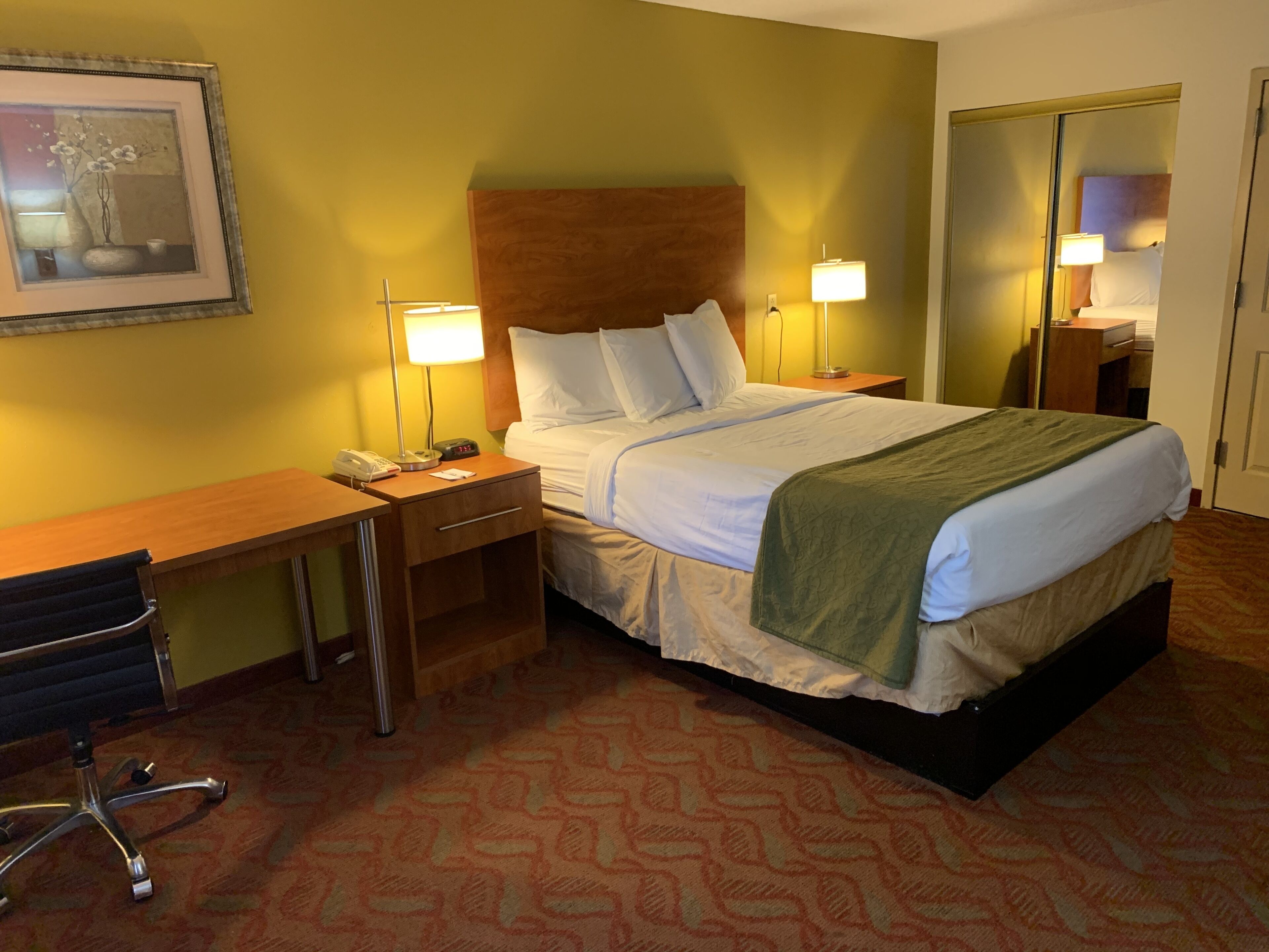 A hotel room with a bed, bedside tables, a desk, a chair, and a framed picture on the wall.