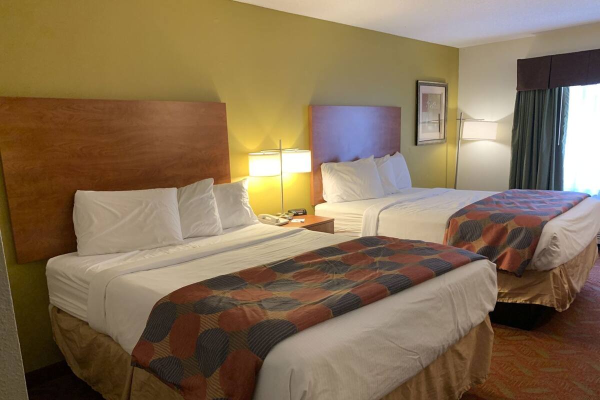 Una habitación de hotel con dos camas, un cabecero de madera, una mesita de noche con lámpara y una ventana con cortinas.