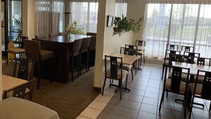 Een restaurantgedeelte in een hotel met een grote centrale tafel, kleinere tafels en stoelen. Er staan potplanten op de tafels en een brandblusser op de vloer.