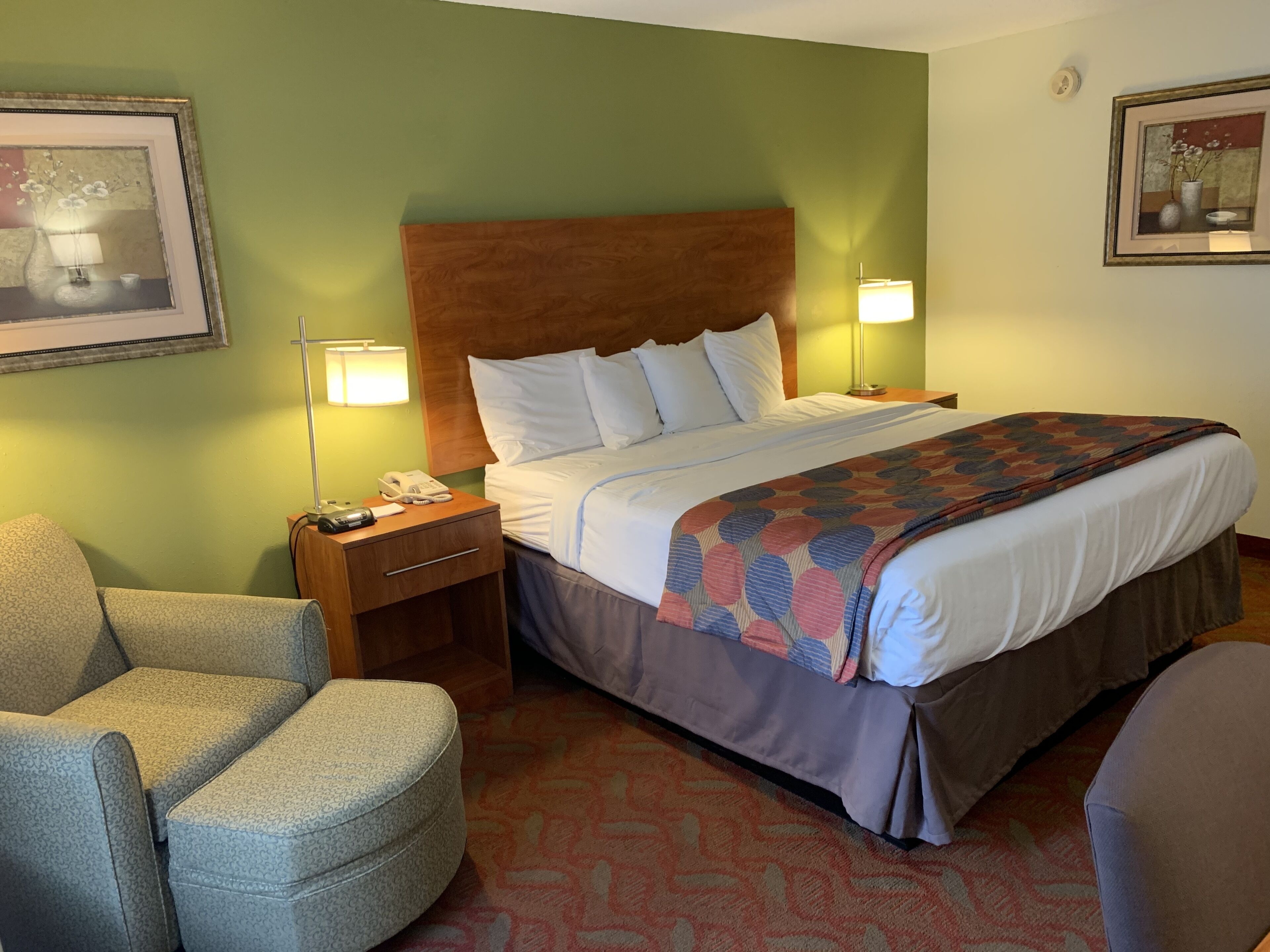 A hotel room with a large bed, a nightstand with a lamp, a chair, and a framed picture on the wall.