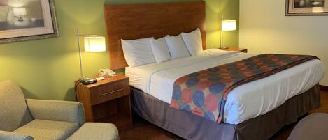 A hotel room with a large bed, a nightstand with a lamp, a chair, and a framed picture on the wall.